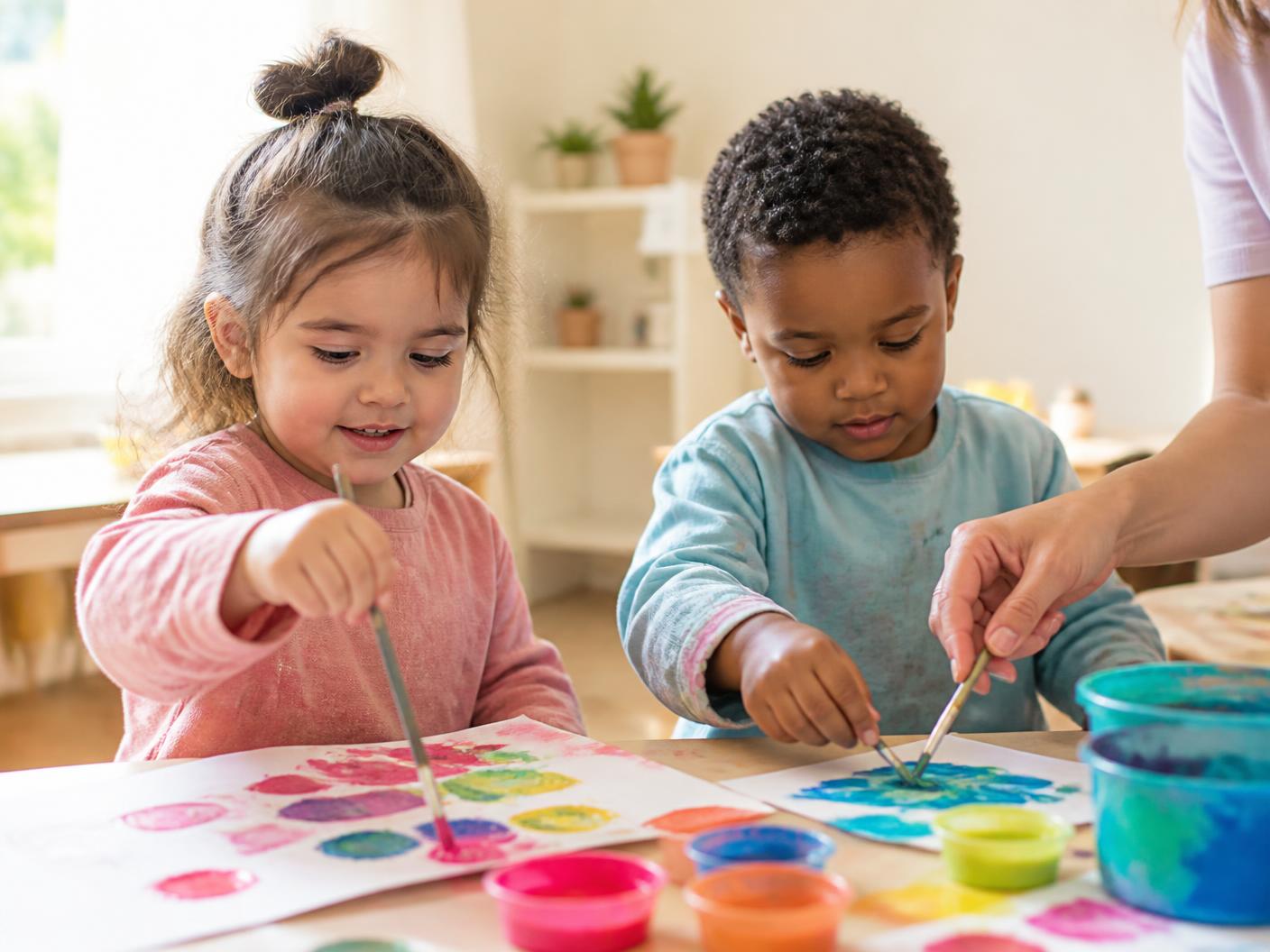 Two young children painting with colorful paints at a low table in a sunlit art corner