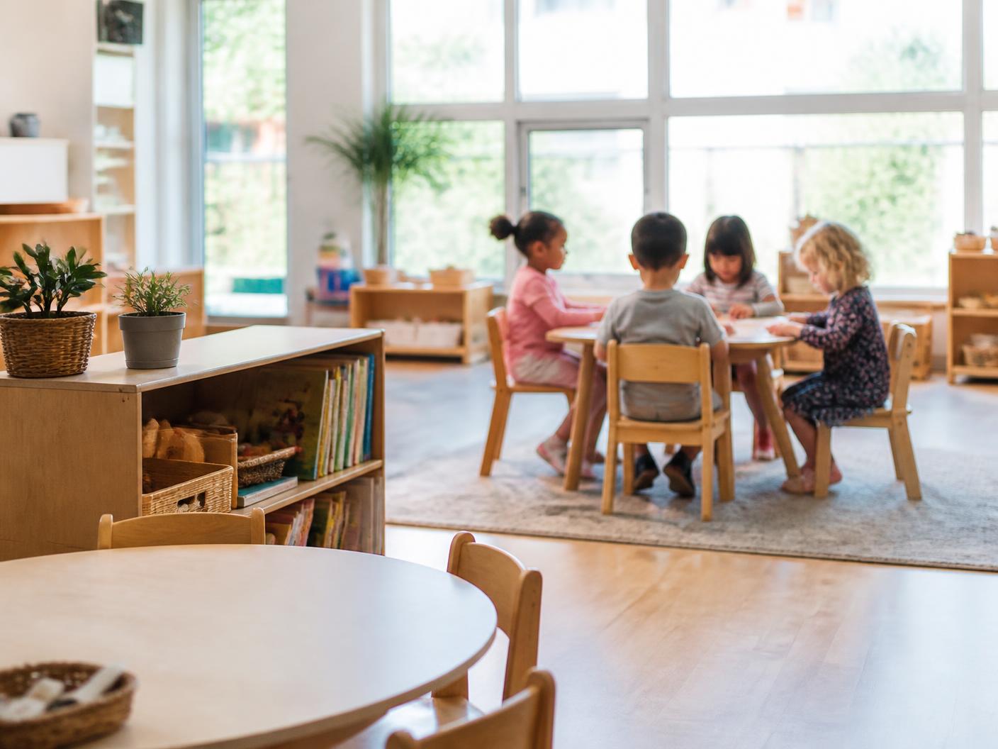 A bright preschool classroom with low wooden tables, book shelves and children working quietly at a table